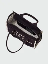 Bolso tote de lona color beige con estampado frontal en negro que dice 'THE TOTE BAG MARC JACOBS'. Presenta bordes laterales en color negro con detalles de texto y doble asa superior.