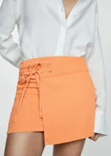 Falda corta de algodón estilo tejano, color naranja, con diseño cruzado y detalle de lazo ajustable con ojales metálicos en la cintura.