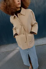 Chaqueta de corte recto y estilo camisero en color beige, con cuello solapa, manga larga y cierre frontal con botones. Presenta dos bolsillos delanteros de plastrón.