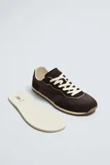 Zapatilla deportiva estilo barefoot, color marrón, con diseño de puntera ancha y redondeada. Confeccionada en una combinación de materiales textiles y detalles en serraje. Cuenta con suela fina de goma flexible y cordones en color crema.