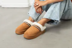 Pantuflas tipo slipper de gamuza color tostado, con interior y borde de peluche sintético blanco. Presentan una suela de plataforma plana y una cinta decorativa con patrón geométrico en el borde superior.