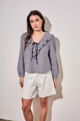 Blusa de corte oversize con estampado de cuadros vichy en blanco y negro. Presenta un cuello con volados, mangas largas con puños elásticos y un cierre frontal con lazo negro.