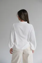 Blusa blanca de corte holgado con cuello mao y cierre frontal de botones. Presenta mangas largas abullonadas con puños elásticos y un delicado bordado en color beige en la parte superior delantera.