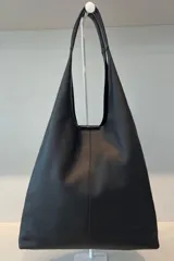 Bolso tipo tote de gran capacidad, confeccionado artesanalmente en cuero vacuno color marrón. Presenta un diseño minimalista con costura central vertical, fuelle lateral ajustable con cierre y placa metálica con logo en la parte inferior.