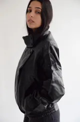 Chaqueta de cuero sintético color negro, con diseño de corte oversize, cuello alto con cierre de botones a presión, hombreras marcadas y ruedo elástico.