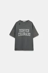Camiseta de corte oversize confeccionada en algodón flamé, con cuello redondo y manga corta. Presenta un diseño con efecto desgastado, detalles de roturas en el tejido y estampado de texto a contraste en la parte frontal.