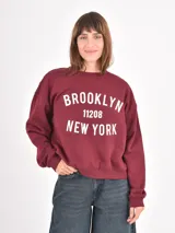 Buzo de cuello redondo en color bordó con estampado frontal en blanco que incluye el texto Brooklyn 11208 New York. Presenta un diseño de corte holgado con hombros caídos y puños acanalados.