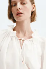 Camisa de lino y viscosa color blanco marfil, de diseño oversized, con cuello alto plisado y volados, mangas largas abullonadas con puños elásticos fruncidos y cordones ajustables en el escote.