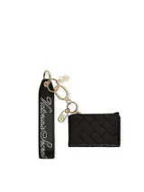 Estuche para tarjetas negro con diseño acolchado y textura tejida, con tarjetero transparente en la parte posterior, cierre con cremallera y herrajes metálicos dorados que incluyen un llavero y dijes con el logo de la marca.