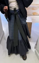 Falda larga tipo patchwork confeccionada en diferentes texturas de tweed a cuadros. Presenta un diseño envolvente con un lazo frontal en color verde oscuro y un ruedo con volados amplios en tela de cuadros más pequeños.