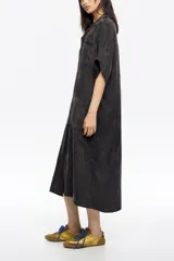 Vestido camisero largo de corte holgado y fluido, con cuello camisero, cierre frontal de botones y mangas cortas. Presenta bolsillos frontales tipo parche en el pecho y a la altura de la cadera.