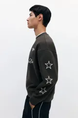 Buzo verde militar de corte oversized, confeccionado en mezcla de algodón con cuello redondo, puños y dobladillo acanalados. Cuenta con estampados en el frente, la espalda y las mangas, incluyendo inscripciones y estrellas.