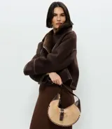 Cartera tipo baguette de diseño curvo, confeccionada en gamuza sintética color beige con detalles en cuero marrón oscuro. Presenta costuras decorativas en los bordes, una correa ajustable y cierre frontal con hebilla metálica dorada.