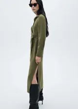 Vestido midi de diseño cruzado en color verde oliva, con cuello camisero, cierre frontal de botones y lazo lateral para ajustar en la cintura. Presenta mangas largas con puños abotonados y una abertura lateral pronunciada en el bajo.