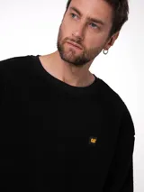 Buzo de cuello redondo confeccionado en tejido sherpa negro, con textura suave y elástica. Presenta puños y banda inferior en tejido acanalado y una pequeña etiqueta con el logo de la marca en el pecho.