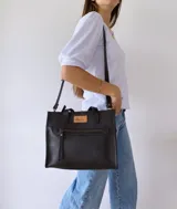Cartera tipo tote bag de cuero color negro, con doble asa corta de mano y correa larga regulable y extraíble para usar al hombro o cruzada. Tiene un bolsillo frontal con cierre y un bolsillo interno.