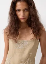 Vestido corto recto en color beige, con tirantes finos y escote recto. Presenta un delicado diseño bordado floral en hilo metalizado y cuenta con un bolsillo frontal superpuesto a la altura de la cadera.