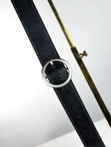Cinto de cuero negro con textura granulada, costuras decorativas en los bordes y hebilla circular metálica en acabado plateado mate.