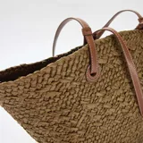 Bolso tipo capazo color kaki, confeccionado en tejido trenzado efecto rafia, con doble asa larga de hombro en cuero sintético marrón claro con costuras blancas visibles.