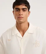 Camisa masculina boxy de manga corta en tricolina color marfil, con cuello cubano, cierre frontal con botones y sutiles bordados veraniegos en tono tostado.