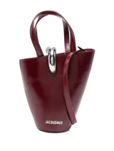 Bolso tipo cubo Jacquemus modelo Le Petit, confeccionado en cuero color bordó con acabado brillante. Cuenta con asas dobles superiores, correa bandolera extraíble, detalle de anilla metálica plateada en la parte frontal y el logotipo de la marca en letras metálicas.