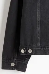 Campera trucker de denim de algodón rígido, color negro lavado, con cuello, botones de metal y canesú delantero y trasero. Posee bolsillos superiores con solapa y botón, bolsillos laterales insertados, puños abotonados y trabilla ajustable con botones en los laterales del bajo.