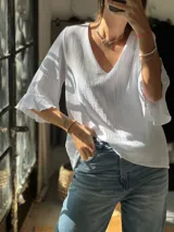 Blusa de muselina blanca con escote en V y mangas cortas con volados en los puños. Presenta una textura arrugada característica del tejido.