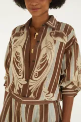Camisa amplia de manga larga con cuello camisero, botonera frontal y hombros caídos. Cuenta con un estampado de rayas verticales y motivos ornamentales con dos aves estilizadas enfrentadas en el frente.