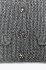 Chaqueta de punto grueso color gris, de cuello redondo y manga tres cuartos. Cierre frontal con botones joya dorados con detalles de pedrería. Cuenta con dos bolsillos frontales tipo parche.