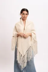 Poncho triangular de lana merino color marfil, confeccionado artesanalmente en telar manual con textura rústica y delicados flecos colgantes en los bordes.