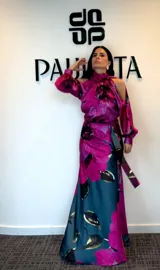 Vestido largo de satén con estampado floral en tonos fucsia, azul petróleo y negro. Presenta cuello alto, diseño con hombros descubiertos y mangas largas con puños. Incluye un lazo ajustable en la cintura para entallar la silueta.