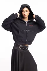 Campera negra de algodón con cierre frontal, capucha, bolsillos laterales y detalles de tachas metálicas en la capucha y los bolsillos. Presenta un estampado frontal con texto en tono rojizo.