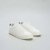 Zapatillas blancas de cuero sintético con cordones, suela plana y detalle de refuerzo negro en el talón.