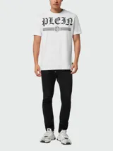 Remera blanca de manga corta y cuello redondo, con estampado frontal en color negro que incluye la palabra "PLEIN" en tipografía gótica y un emblema decorativo con líneas horizontales.