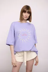 Remera lila de corte holgado con mangas cortas y estampado circular con figuras de nadadoras y texto "The Ped Swim Club".