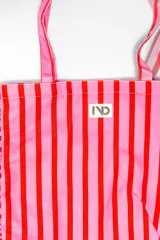 Bolso tipo tote bag de tela con diseño de rayas verticales en tonos beige y gris, decorado con volados en los laterales y la base. Incluye dos asas largas para llevar al hombro y una etiqueta con logo en el frente.