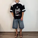 Camiseta estilo fútbol americano de corte oversize, color negro con detalles en blanco y bordó en el cuello y mangas. Presenta un estampado frontal con tipografía desgastada, una estrella y un escudo.