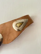 Cinto tipo faja de cuero color marrón, con diseño de piezas de cuero unidas y apliques metálicos plateados grabados con detalles geométricos y una piedra central oscura.
