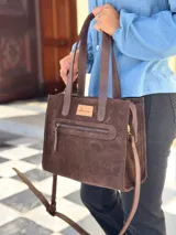 Cartera tipo tote de gamuza color chocolate, con doble asa de mano y correa larga ajustable y extraíble. Presenta un bolsillo frontal con cierre metálico y una etiqueta de cuero con el logo de la marca en el frente.
