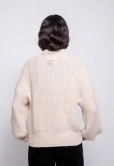 Sweater color crema de tejido trenzado con cuello alto, mangas largas abullonadas y detalle de flecos en el frente.