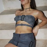 Minifalda negra de cuero sintético con cierre frontal con cremallera y cintas cruzadas con hebillas metálicas.