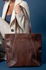 Bolso tipo shopper de cuero color suela con textura grabada, detalle de costuras laterales trenzadas y placa metálica con logo de la marca. Cuenta con doble asa larga para colgar al hombro, cierre principal y múltiples bolsillos internos y externos.