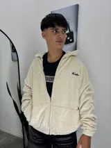 Campera con cierre frontal, confeccionada en corderoy de textura suave y acabado aterciopelado en color crema. Presenta capucha, bolsillos laterales y un pequeño bordado decorativo en el pecho.
