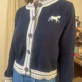 Saco de punto tejido en color azul marino con detalles en color crema en los bordes, puños y cuello. Presenta un diseño de caballo bordado en el pecho, cierre frontal con botones oscuros y un corte clásico.