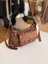 Cartera tipo baguette con estampado animal print, confeccionada en material suave símil gamuza. Presenta un bolsillo frontal con solapa y cierre, compartimento principal con cierre superior, y asa larga regulable de cuero sintético marrón.