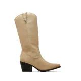 Bota texana de caña alta confeccionada en gamuza color beige, con taco cuadrado oscuro y punta fina. Presenta detalle de costuras decorativas y aplique metálico con la letra V en la parte superior de la caña.