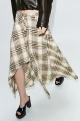 Falda midi de tiro alto con estampado de cuadros en tonos beige, marrón y negro. Presenta un diseño asimétrico con abertura frontal y detalles desflecados en el bajo.