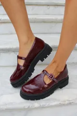 Zapato tipo guillermina en cuero charol color bordó, con suela track negra de goma, cierre con hebilla ajustable y detalle de herrajes metálicos dorados.