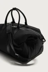 Bolso de viaje tipo bowling fabricado en combinación de nylon y detalles en cuero sintético, de color negro. Cuenta con compartimento principal con cierre de cremallera, bolsillo frontal plano, dos asas de mano y un asa de hombro ajustable y extraíble.