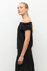 Blusa negra estilo bardot con escote de hombros descubiertos, detalles de volados fruncidos en el pecho y diseño sin mangas.